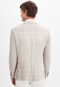 Mann von hinten zu sehen, der ein beiges Blazer mit einem weißen Fensterrahmenmuster und weiße Hosen trägt, vor einem einfarbigen Hintergrund.