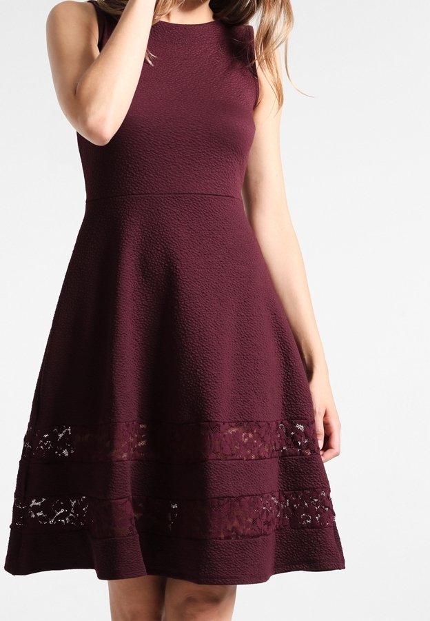 Femme portant une robe sans manches texturée de couleur violet foncé avec des panneaux en dentelle près de l'ourlet, debout devant un fond clair uni.