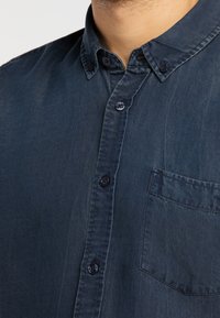 Ciemnoniebieska koszula na guziki z klasycznym kołnierzem, wykonana z gładkiej, lekko teksturowanej tkaniny, z jedną kieszenią na piersi.