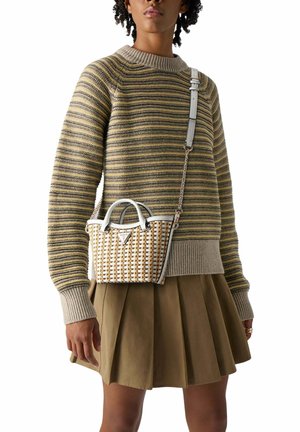 Personne portant un pull rayé beige et jaune avec des poignets côtelés, une jupe plissée marron et un sac bandoulière tissé blanc et beige.