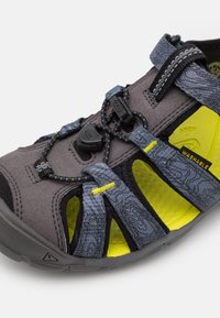 Grå og svart åpen tå sandal med teksturert overdel, gule detaljer, justerbare bungee-snører og en polstret, vaskbar fotseng.