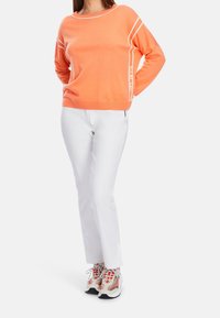 Pull en maille orange avec accents blancs, associé à un pantalon blanc et des baskets de sport multicolores avec des motifs et des lacets.