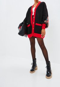 Cárdigan oversized negro y rojo con mangas largas, bolsillos frontales y acentos decorativos con patrones. Combinado con un vestido negro ajustado y botas de tobillo.