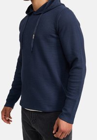 Sudadera de rayas en color marino con capucha y cordón, detalles de costura lateral y un logo sutil en el pecho, fabricada con un tejido suave y texturizado.