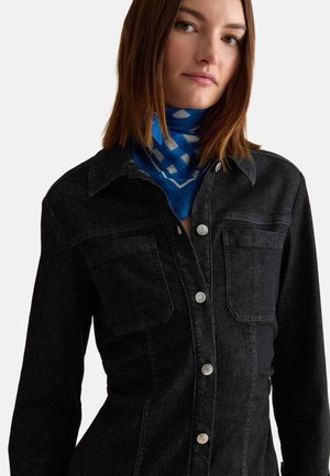 Jeune femme portant une veste en jean noire avec des boutons argentés et un foulard bleu à motifs autour du cou, face à l'avant sur un fond uni.