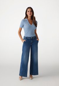Hellblaues, geripptes Kurzarm-Oberteil mit V-Ausschnitt und zwei Brusttaschen, kombiniert mit weit geschnittenen, dunklen Jeans und beigen High Heels.