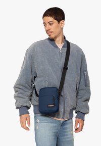 Námořnická modrá taška na přes rameno Eastpak s přední kapsou na zip a nastavitelným popruhem, vyrobená z odolné tkaniny s hladkou texturou.