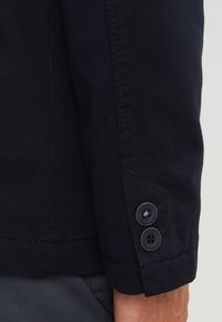 Granatowa marynarka o gładkiej fakturze materiału, z trzema brązowymi guzikami na mankietach oraz dopasowanym ściegem wzdłuż szwów.