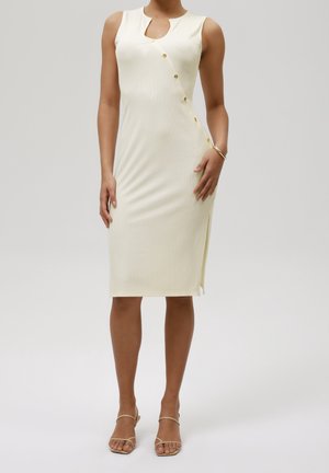 Mouwloze crèmekleurige jurk tot op de knie met asymmetrische gouden knopen, een halslijn met inkeping, een zijsplit en beige sandalen met bandjes op het model.
