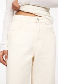 Jeans crème taille haute avec une coupe droite, fermeture à bouton et des détails de couture subtils. Le tissu semble doux avec une texture lisse.