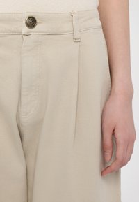 Beige pants made of denim, featuring a pleated front, a gold button, and subtle stitching details. Hand shown holding fabric at the side.