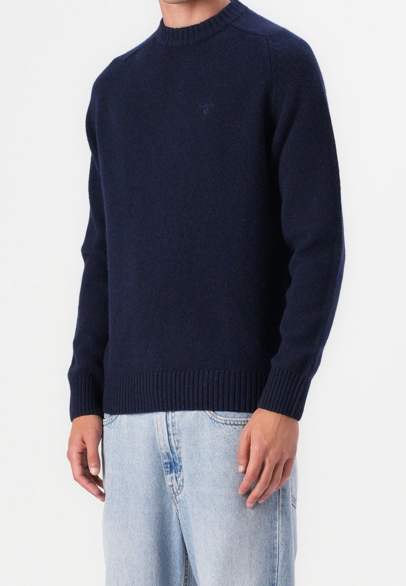 Homme portant un pull bleu marine à col rond avec poignets et bas côtelés, associé à un jean en denim bleu clair, debout devant un fond blanc.