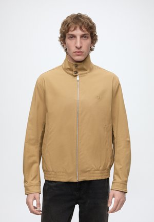 Junger Mann mit kurzen lockigen Haaren, der eine beige Jacke mit Reißverschluss und hohem Kragen sowie schwarze Hose trägt, steht vor einem schlichten hellen Hintergrund.