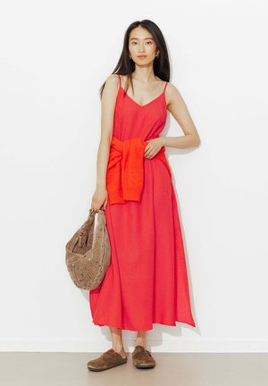 Femme portant une robe maxi rouge sans manches avec un pull rouge noué à la taille, tenant un sac en suède marron et portant des chaussures marron sans lacets.