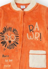 Bata de forro polar naranja con diseño de león, detalles en negro, cuello crema y un bolsillo blanco. El texto incluye "Nacido para ser adorable" y "¡RAWR!"