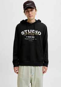 Musthave musta kapuutsiga, millel on valge ja kuldne trükitud tekst; omab eesmist taskut ja nööri. Kombineeritud heledate teksade ja pruuni mütsiga.