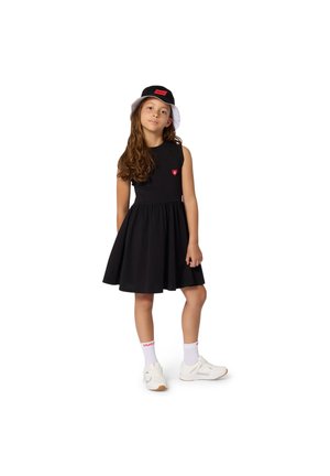Robe noire sans manches avec une jupe évasée et un écusson en forme de cœur, associée à un chapeau seau noir et blanc et des baskets blanches avec des accents rouges.