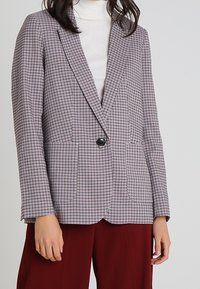 Šedý kostkovaný blazer s klopami, strukturovaný tvar, jeden černý knoflík a dvě přední kapsy, v kombinaci s bílým rolákem.