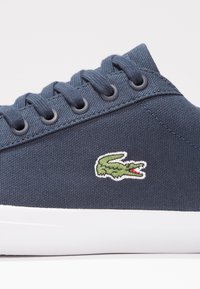Baskets bleu marine avec dessus en tissu, lacets assortis, semelle blanche et logo crocodile vert brodé sur le côté.
