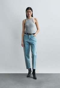 Γυναίκα με μακριά σκούρα μαλλιά, φορώντας γκρι αμάνικη μπλούζα, ανοιχτό μπλε ψηλόμεσο τζιν, μαύρη ζώνη και μαύρα μπότες μέχρι τον αστράγαλο, στέκεται μπροστά σε έναν μονόχρωμο τοίχο.