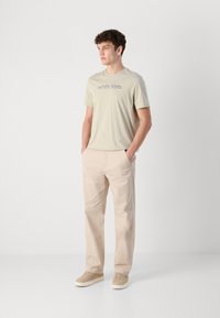 Lys grå t-skjorte med korte ermer og minimalistisk logo, kombinert med beige chinos og beige joggesko. Glatt stoff med avslappet passform.
