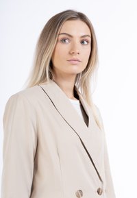 Beige, dupla soros blézer, testre szabott fazonnal, bemetszett hajtókával és két nagy, kerek gombbal. Simább anyag, finom fényességgel.