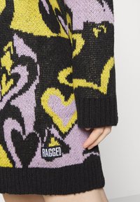 Maglione lavorato a maglia con audaci motivi neri, lavanda e gialli, con orlo nero a costine e un'etichetta "RAGGED" visibile in basso.