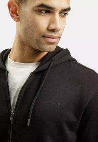 Czarna bluza z zamkiem wykonaną z miękkiego materiału, z szarym wykończeniem wewnętrznym oraz subtelnym logo na klatce piersiowej. Dopasowana, ale o swobodnym kroju.