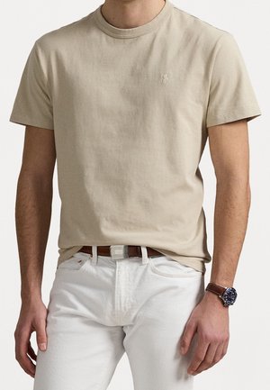 Homme portant un t-shirt beige clair à manches courtes, un pantalon blanc avec une ceinture marron et une montre-bracelet avec un bracelet en cuir marron au poignet gauche.