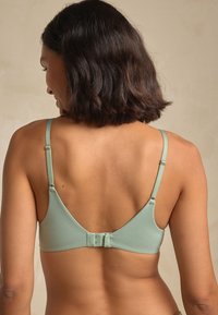 Mėtų žalios spalvos belaidis liemenėlė su reguliuojamomis petnešėlėmis ir užsegimu nugaroje, pasižyminti lygia tekstūra ir minimalistiniu dizainu.