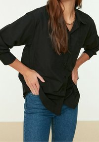 Personne portant une chemise noire ample boutonnée et un jean bleu, les mains posées sur les hanches, avec de longs cheveux bruns et un collier chaîne en or.