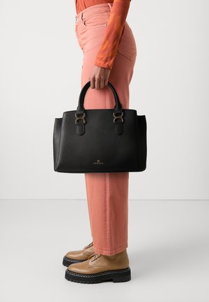 Personne portant un haut orange et un pantalon corail, tenant un sac à main en cuir noir avec des boucles dorées et des bottines à plateforme beige, sur un fond uni.