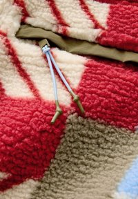 Tissu polaire texturé avec des motifs rouges, beiges et blancs et un gros plan sur des cordons bleus avec des embouts en plastique verts attachés à un tissu vert olive.