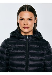 Sieviete ar taisniem tumšiem matiem valkā melnu rievotu žaketi ar rāvējslēdzēju un kapuci, skatoties tieši uz priekšu pret vienveidīgu gaišu fonu.