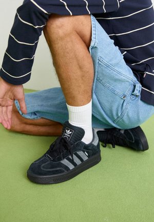 Black suede sneakers with three gray stripes, textured accents, black rubber sole, worn with white socks and light blue denim shorts.