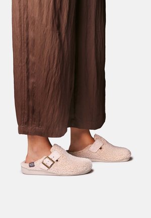 Pieds portants des chaussons beige duveteux à enfiler avec une boucle, associés à un pantalon marron à jambes larges sur un fond uni.
