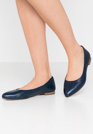 Des pieds portant des ballerines en cuir bleu marine avec de petits talons en bois sur un fond blanc.