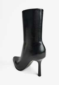 Černá kotníková bota z kůže se špi�čatou špičkou a úzkým vysokým podpatkem, zobrazená zezadu na bílém pozadí.