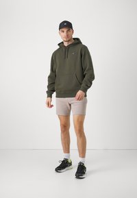 Felpa verde oliva con lacci e tasca frontale, abbinata a pantaloni corti beige e sneakers nere con accenti neon. Cappellino nero in testa.
