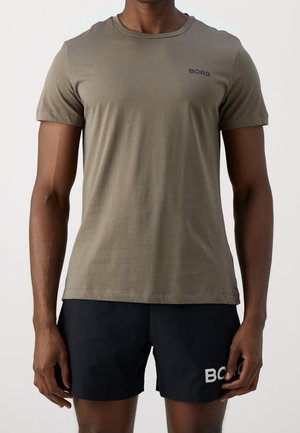 Homme portant un t-shirt à manches courtes vert olive et un short noir avec un logo sur la poitrine et la jambe, debout devant un fond uni.