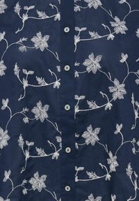 Tumši zila auduma ar baltu ziedu izšuvumu un rindā mazi apaļi pogas vidū.