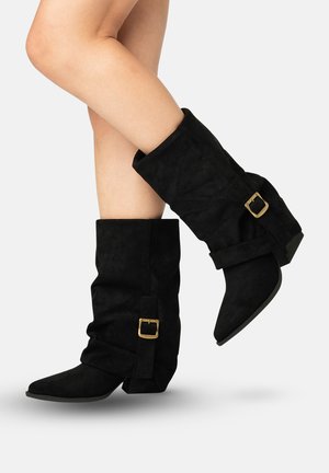 Nogi w czarnych zamszowych botkach do połowy łydki z niskimi obcasami, podwiniętymi cholewkami i złotymi paskami zapinanymi na klamry na jednolitym białym tle.