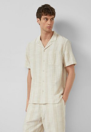 Jonge man draagt een beige met witte gestreepte korte mouwen blouse met knopen en bijpassende korte broek, staat met één hand in zijn zak en kijkt naar rechts.