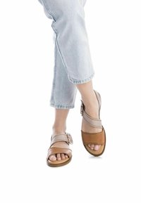 Sandalias marrones y beige con un diseño de tiras anchas, detalle de hebilla y punta abierta. Colocadas sobre unos jeans de mezclilla claros y desgastados.
