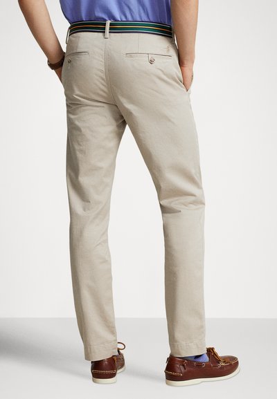 Beżowe bawełniane chinosy o wąskim kroju, z tylnymi kieszeniami i dekoracyjnym paskiem w paski w kolorach zielonym, żółtym i niebieskim. Widoczne brązowe mokasyny.