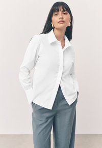 Chemise blanche à boutons avec col italien, manches longues et texture lisse, associée à un pantalon gris clair.