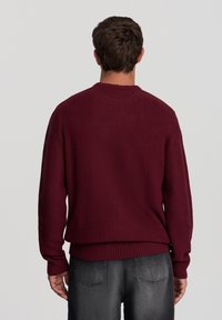 Maglione a maglia bordeaux con collo e polsini a costine. Vestibilità rilassata con motivo testurizzato. Abbinato a jeans grigio scuro, vista da dietro.