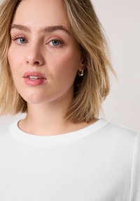 Young woman with short blonde hair wearing silver hoop earrings and a white crew-neck shirt against a plain light background.