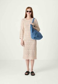 Vestido de punto beige con un patrón de encaje, mangas 3/4 y dobladillo con picos. Bolsa de tela azul y sandalias negras. Modelo de pie contra un fondo sencillo.