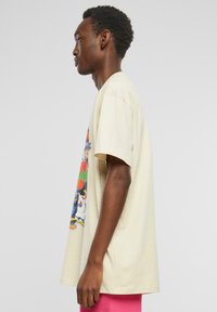 T-shirt beige à manches courtes en coton, avec un imprimé graphique coloré sur le côté gauche. Coupe ample et design décontracté.